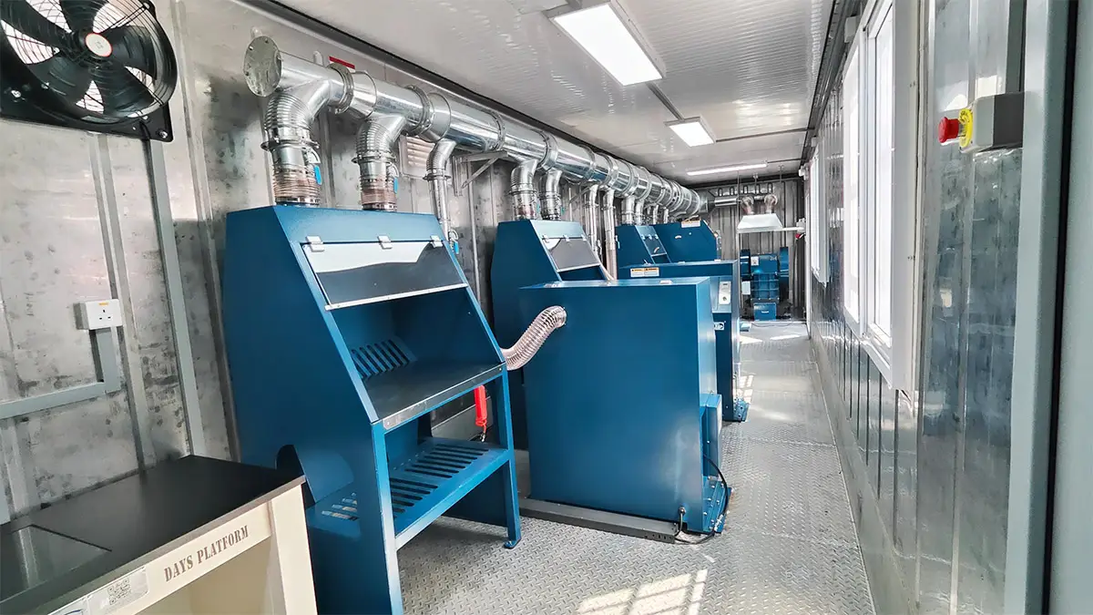 Interior view of Tanzania's mobile containerized sample preparation lab, featuring a row of blue industrial sample preparation machines with extensive overhead ventilation ducts and a large wall-mounted fan.