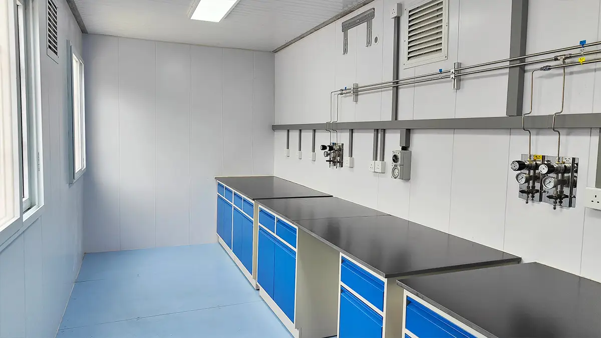 Interior of a DECENT Graphite AAS Analytical Hub, showing a leftward view of the sample preparation area with lab benches, blue cabinets, gas lines, and windows.