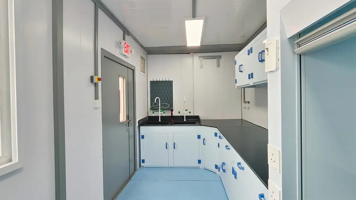 Interior of a DECENT Graphite Chemical Analysis Laboratory, showing a leftward view of the sample pretreatment area with a double sink, lab benches, blue and white cabinetry, and an emergency exit door.