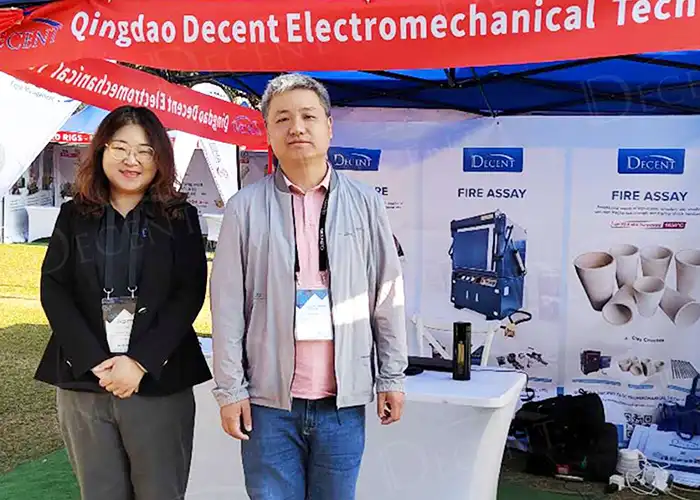 Representatives from Qingdao Decent Group at DRC Mining Week, standing in front of their booth showcasing fire assay equipment and promoting mining sustainability.