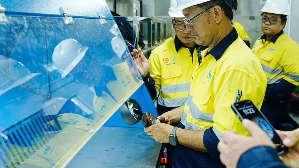 Lynas engineers in yellow safety vests and hard hats inspecting blue laboratory equipment, possibly a sample preparation machine, within the DECENT Malaysia Container Laboratory Project 2024.