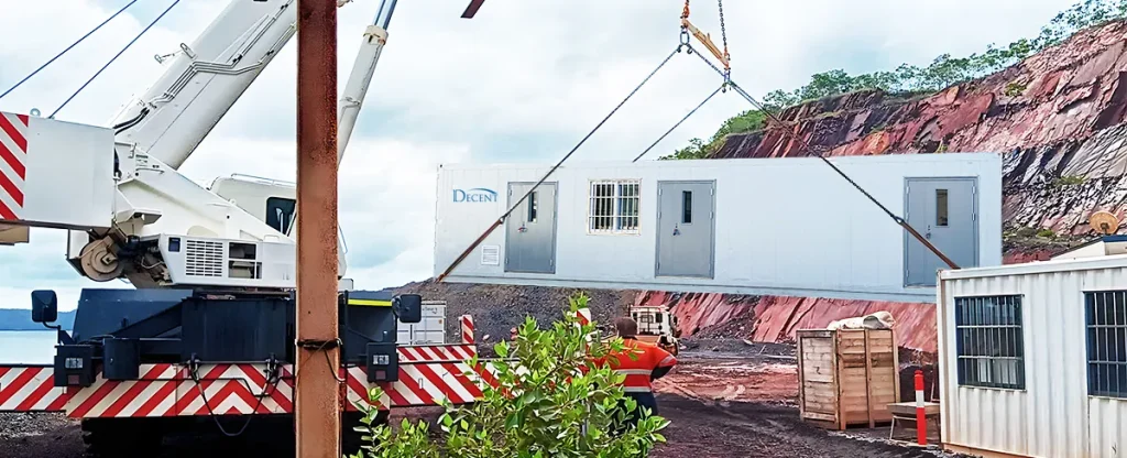 A white DECENT mobile containerized laboratory being lifted by a large crane in a rugged outdoor environment, with a red rock face in the background.