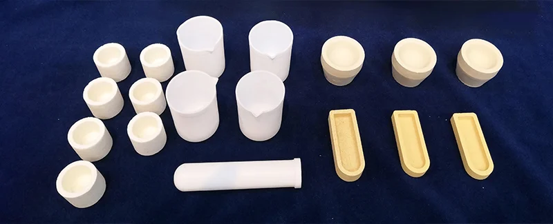 A collection of various ceramic laboratory crucibles and molds, including cylindrical crucibles with pouring spouts, smaller round crucibles, and rectangular assay molds, displayed on a dark blue surface