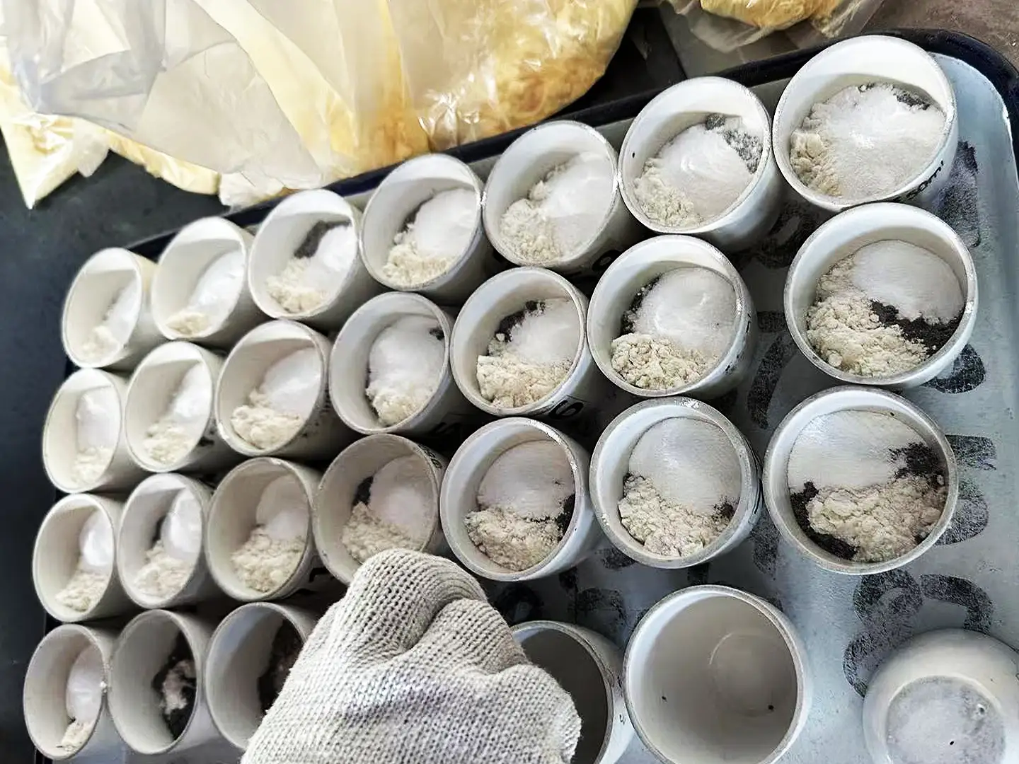 A tray of white ceramic crucibles containing various powdered samples and fluxes, being prepared by a gloved hand for laboratory analysis, illustrating Qingdao Decent Group's sample preparation process.