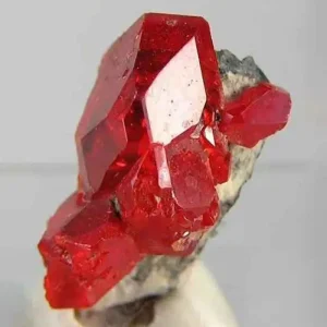 A cluster of brilliant, translucent red realgar crystals, a toxic arsenic sulfide mineral, on a light gray matrix.