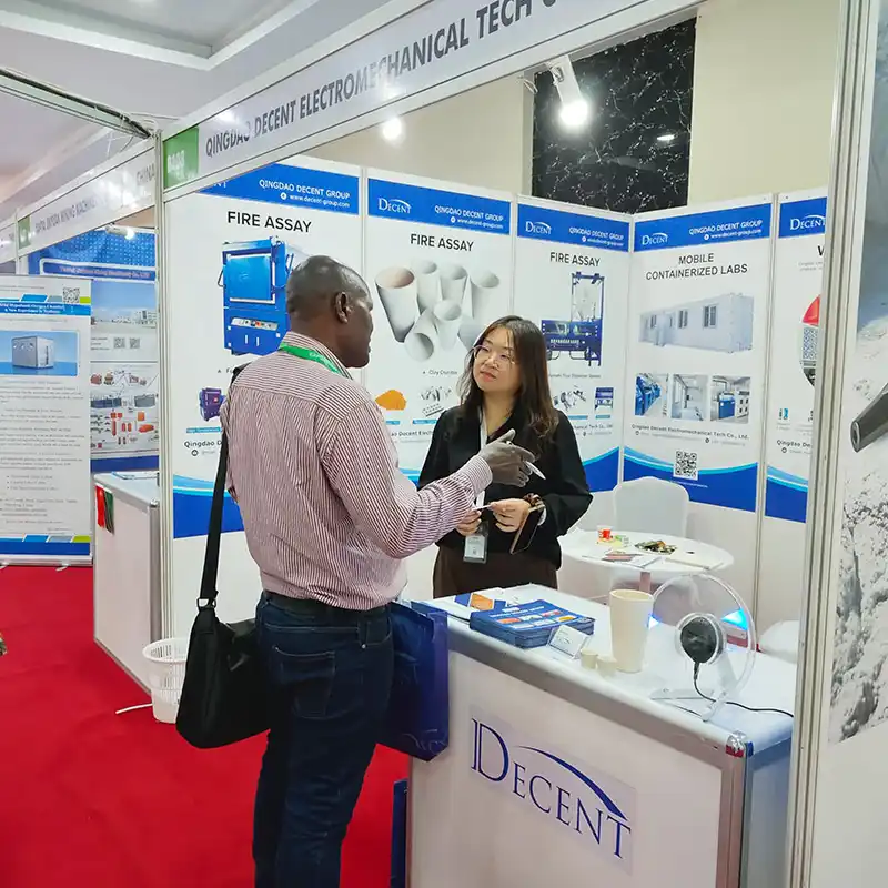 Qingdao Decent Group representative speaking with a male customer at the DECENT booth at Minexpo Tanzania 2025, discussing mineral assay equipment and laboratory solutions.